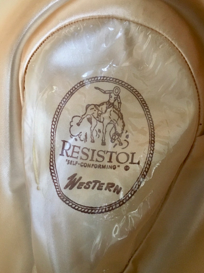 May include: A brown and white Resistol Western hat with a cowboy riding a horse logo. The hat is labeled "Self-Conforming".