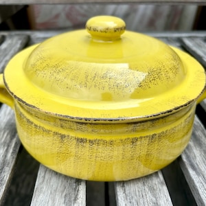 Peut inclure: Un plat à casserole en céramique jaune avec un couvercle. Le plat a deux poignées et un petit bouton sur le couvercle. L'extérieur a une finition vieillie avec des accents sombres. Le plat est posé sur une surface en bois.