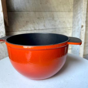 Pode incluir: Uma panela de ferro fundido esmaltada laranja com duas alças pretas. A panela é redonda e tem um acabamento brilhante.