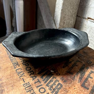 May include: A black, oval-shaped bowl with a slightly raised rim and angular handles. The bowl is made of a dark material and sits on a wooden surface with printed text. The background includes wooden beams and architectural elements.