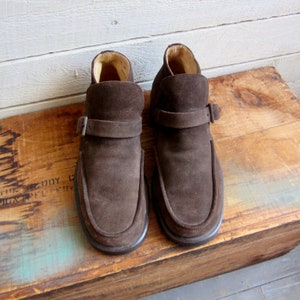 May include: A pair of brown suede ankle boots with a buckle strap. The boots have a rounded toe and a low heel.