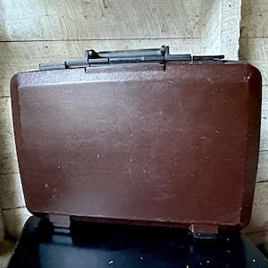 May include: A vintage brown suitcase with a black handle and latch. The rectangular case has a slightly textured surface and sits on a black surface. The suitcase is a classic travel accessory.