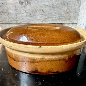 May include: A round, glazed ceramic casserole dish with a matching lid. The exterior features a two-tone design with a dark brown base that transitions into a lighter tan color toward the rim. The lid has a smooth, glossy brown finish.