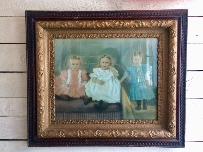 May include: An antique framed photograph of three young girls in white, pink, and blue dresses. The girls are sitting on a couch and smiling at the camera. The frame is ornate with gold accents.