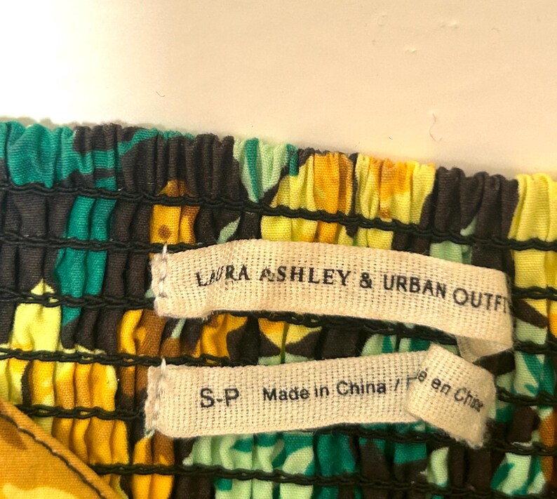 May include: Close-up of a colorful floral fabric with the labels "LAURA ASHLEY & URBAN OUTFITTERS" and "S-P Made in China / Fait en Chine". The fabric features a pattern of yellow, green, and black.