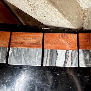 May include: Four rectangular coasters with a two-tone design. The top half is a warm, reddish-brown wood, while the bottom half is a gray and white marbled stone. The coasters are arranged in a row.