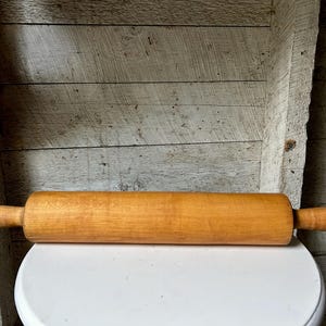 May include: A cylindrical wooden rolling pin with a smooth, light brown finish and two rounded handles on either end. The kitchen tool features a consistent grain pattern throughout its length and is positioned horizontally across the frame.