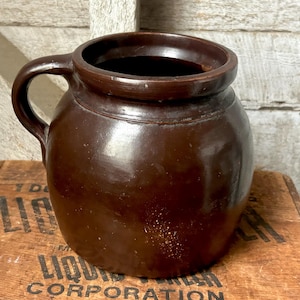 May include: A brown, antique stoneware crock with a handle. The crock has a rounded body and a dark interior. It sits on a wooden crate with faded lettering. The crock is likely used for food storage.