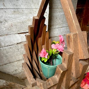 Puede incluir: Un macetero colgante de madera con un diseño geométrico único. El macetero contiene una pequeña maceta turquesa con flores rosas y hojas verdes. La madera tiene un aspecto natural y envejecido.