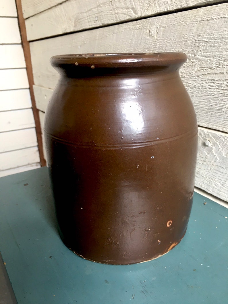 Puede incluir: Un tarro de barro cocido esmaltado marr&oacute;n con una boca ancha y un cuerpo redondeado. El tarro tiene un ligero brillo y algunas imperfecciones visibles.
