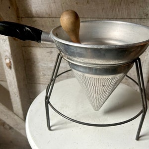 Può includere: Un colino in alluminio vintage con un pestello in legno, appoggiato su un supporto in metallo nero. Il colino ha una forma conica con un fondo a rete e una parte superiore larga e poco profonda.