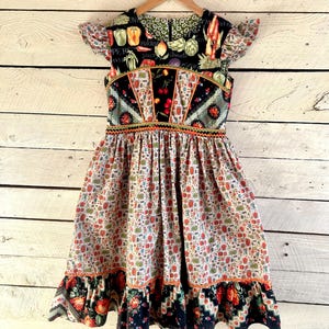 以下が含まれることがあります： 野菜と果物を描いたカラフルな花柄の黒と白のドレス。ドレスは体にフィットするボディスと、フリルトリムのついたボリュームのあるスカート。ドレスはフリル付きの短い袖です。