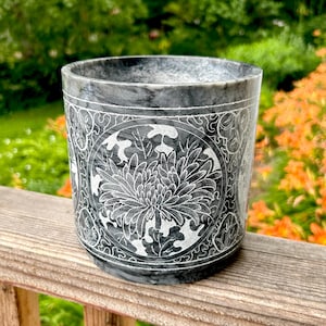 May include: A grey marble container with a detailed floral design. The container features a large, central flower surrounded by intricate patterns. The background shows a blurred green and orange outdoor scene.