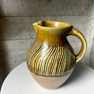 May include: A brown and yellow ceramic pitcher with a striped design. The pitcher has a wide mouth and a handle.