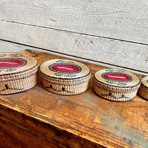 May include: A set of four round, woven lidded baskets of varying sizes, featuring a natural tan base with decorative patterns in red, green, and dark brown. Each basket has a flat lid with a central red oval and geometric border designs.
