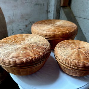 May include: Three round, woven baskets with lids. The baskets are made of light brown wicker and have a circular design on the lids. They are arranged on a white surface, with a rustic wooden background.