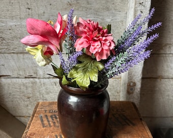 Könnte beinhalten: Eine dunkelbraune Keramikvase gefüllt mit einem farbenfrohen Arrangement aus künstlichen Blumen. Das Arrangement umfasst rosa, rote und lila Blüten, mit grünen Blättern. Die Vase steht auf einer Holzkiste mit Text.