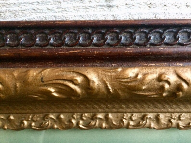 May include: Close-up of a gold-colored ornate picture frame with a dark brown wood inner frame. The frame has a decorative, intricate design.