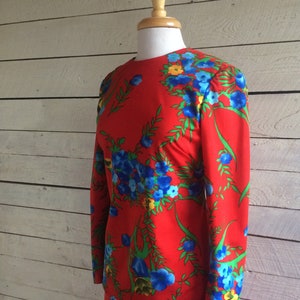 Op de afbeelding: Een rode jurk met een bloemenprint met blauwe, gele en groene bloemen. De jurk heeft lange mouwen en een ronde hals.