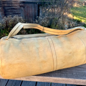 May include: A cylindrical, tan leather duffel bag with a zipper closure and a single handle. The bag shows signs of wear, with darker stitching and a weathered appearance. The bag is resting on a wooden surface.