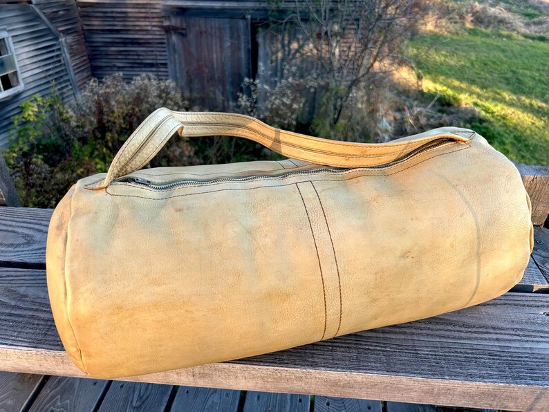 May include: A cylindrical, tan leather duffel bag with a zipper closure and a single handle. The bag shows signs of wear, with darker stitching and a weathered appearance. The bag is resting on a wooden surface.