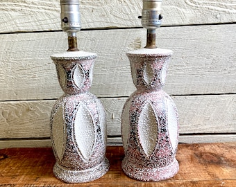 May include: A pair of ceramic table lamps with a pink and white speckled glaze. The lamps have a geometric design with a white background and pink accents. The lamps are in a vintage style.