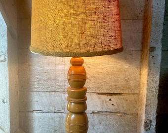 May include: A wooden table lamp with a stacked, turned base and a burlap lampshade. The lampshade is a warm, golden brown color with a red lining.