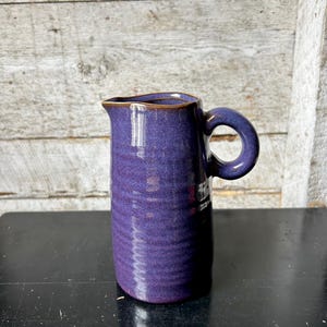 May include: A tall, cylindrical purple ceramic pitcher with a rounded handle. The pitcher has a textured surface and a brown rim. The background is a weathered wooden wall.