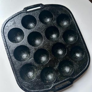 May include: Black cast iron takoyaki pan with 12 round moulds. The pan has two small handles and is designed for cooking small, spherical Japanese snacks. The pan is sitting on a white surface.