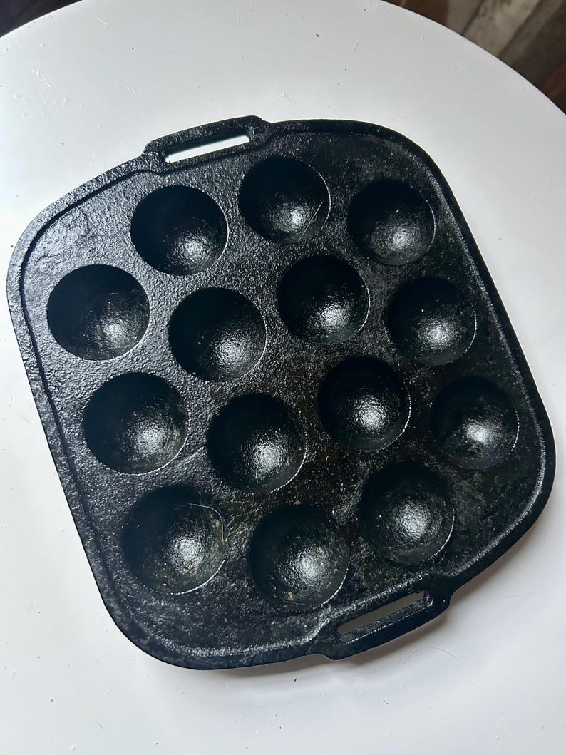 May include: Black cast iron takoyaki pan with 12 round moulds. The pan has two small handles and is designed for cooking small, spherical Japanese snacks. The pan is sitting on a white surface.