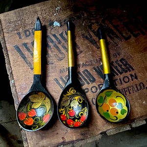 Puede incluir: Tres cucharas de madera pintadas a mano con cuencos negros y mangos amarillos. Las cucharas presentan intrincados diseños florales con bayas rojas y naranjas y hojas verdes. Las cucharas se exhiben sobre una superficie de madera.