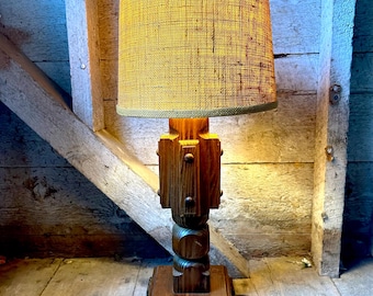 May include: A wooden table lamp with a brown, woven lampshade. The lamp base is made of wood and has a unique, textured design.