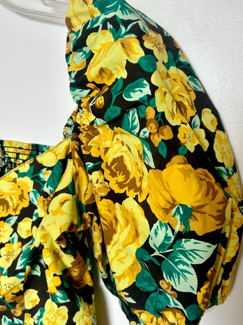 May include: Close-up of a floral print top featuring large yellow roses, green leaves, and brown accents on a black background. The top has a square neckline and puffed sleeves, with elastic detailing.