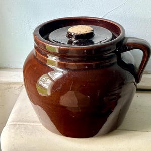 Può includere: Un vaso di ceramica marrone con coperchio e manico. Il vaso ha una finitura lucida e un tappo di sughero. Il manico è curvo e attaccato al lato. Il vaso è su una superficie bianca.