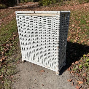 May include: A rectangular, white wicker laundry hamper with a slightly distressed finish. The hamper has a woven design, four small feet, and a hinged lid. It is placed on a concrete surface with a natural background.