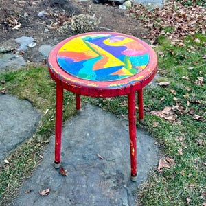 May include: A red metal stool with three legs. The top of the stool is painted with a colorful abstract design.