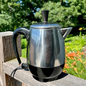 Puede incluir: Una cafetera eléctrica de percolador de acero inoxidable con cuerpo de metal plateado pulido, base de plástico negro, asa negra a juego y un pomo negro en la tapa. Un cable de alimentación negro se extiende desde la base de la cafetera.