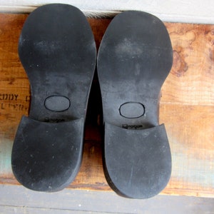 May include: Close-up of the soles of two black leather shoes. The shoes have a brown leather heel and a rubber sole with the text "Hush Puppies" printed on the bottom.