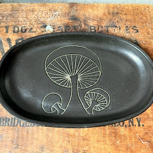 May include: An oval-shaped black ceramic tray featuring a light-colored line drawing of three mushrooms in the center. The tray has a smooth, matte finish and a slightly raised rim, providing a simple and functional surface for holding small items.