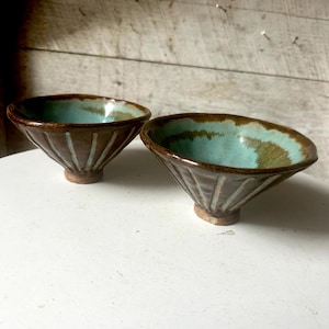 May include: Two small, brown and teal ceramic bowls with a textured, striped design. The bowls are sitting on a white surface.