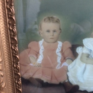 May include: A vintage portrait of a young girl wearing a pink dress with white lace trim. The girl is sitting in a chair and looking directly at the camera. The portrait is framed in a gold frame.