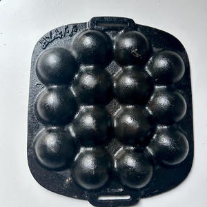 May include: Black cast iron aebleskiver pan with 14 spherical moulds. The pan is rectangular with a handle at each end. The moulds are arranged in a grid pattern. Aebleskivers are small Danish pancakes.