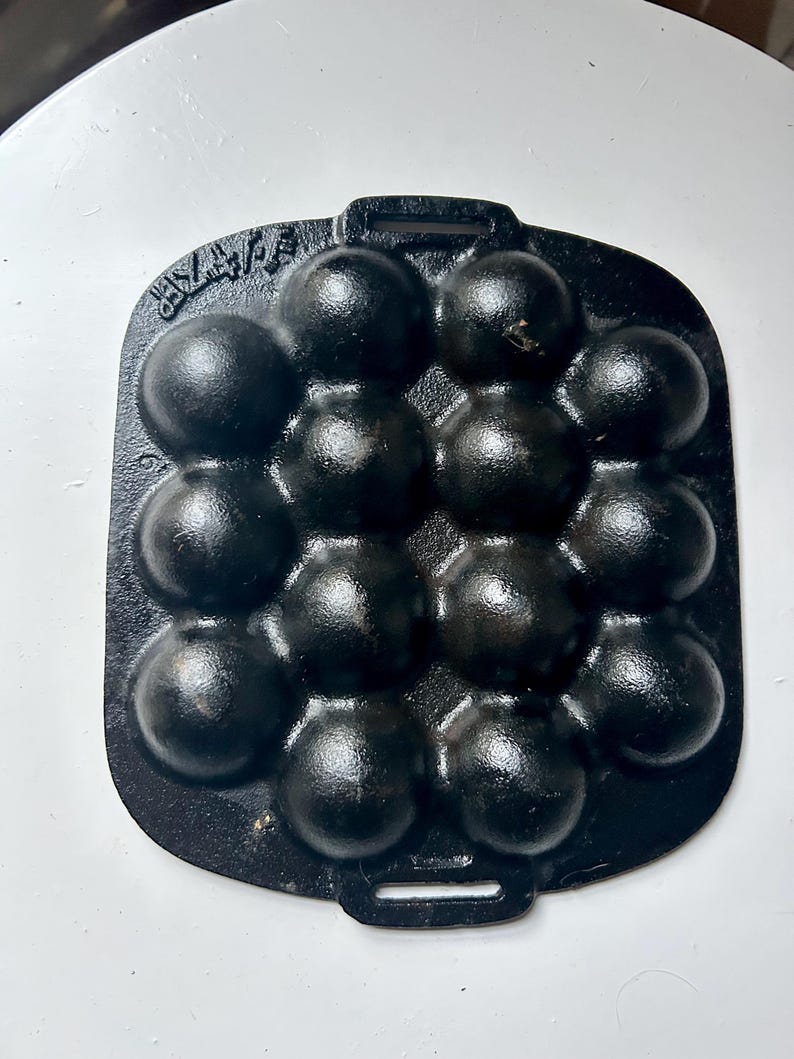 May include: Black cast iron aebleskiver pan with 14 spherical moulds. The pan is rectangular with a handle at each end. The moulds are arranged in a grid pattern. Aebleskivers are small Danish pancakes.
