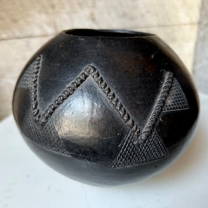 Puede incluir: Una olla de cerámica negra con un diseño geométrico. La olla es redonda y tiene una abertura ancha. El diseño está formado por triángulos y líneas.