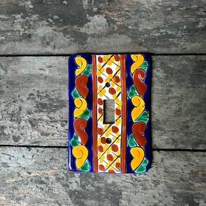 以下が含まれることがあります： 青色の背景に、赤、黄色、緑のアクセントが施されたカラフルなセラミック製ライトスイッチカバー。白と黄色の幾何学模様が描かれています。