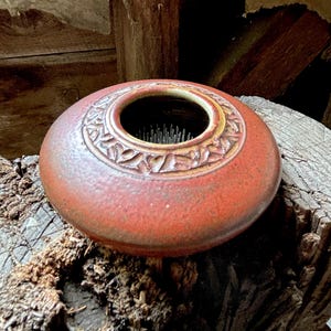 May include: A round, low-profile ceramic bowl with a textured, reddish-brown glaze. The top rim features a carved, repeating geometric pattern in a lighter tan tone. Inside the center opening, a metal flower frog with sharp, upright pins is visible.