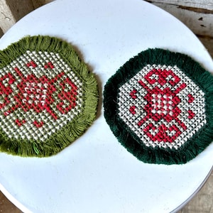 May include: Two round, hand-stitched coasters. One has a green fringe and red text on a white and green checkered background. The other has a dark green fringe and a red geometric design on a white and green checkered background.