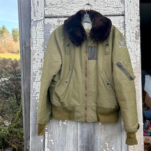 Puede incluir: Una chaqueta bomber verde con cuello de piel sintética marrón. La chaqueta tiene cierre de cremallera y dos bolsillos en la parte delantera. La chaqueta tiene un parche en la manga izquierda que dice "USAF" en letras blancas.