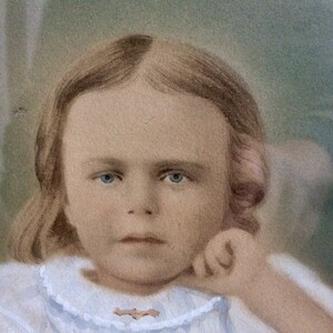 May include: A vintage portrait of a young girl with light brown hair, wearing a white dress with a lace collar. She has blue eyes and a serious expression. The photo is faded and has a soft, dreamy quality.