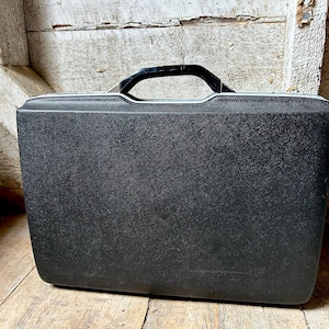 May include: A black vintage briefcase with a textured surface and a black handle. The briefcase is rectangular and has a silver trim. The background is a rustic wooden wall and floor.